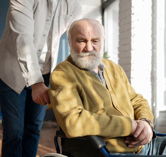 Starszy siwowłosy mężczyzna ze smutną miną siedzi na wózku inwalidzkim pchanym przez młodego mężczyznę.